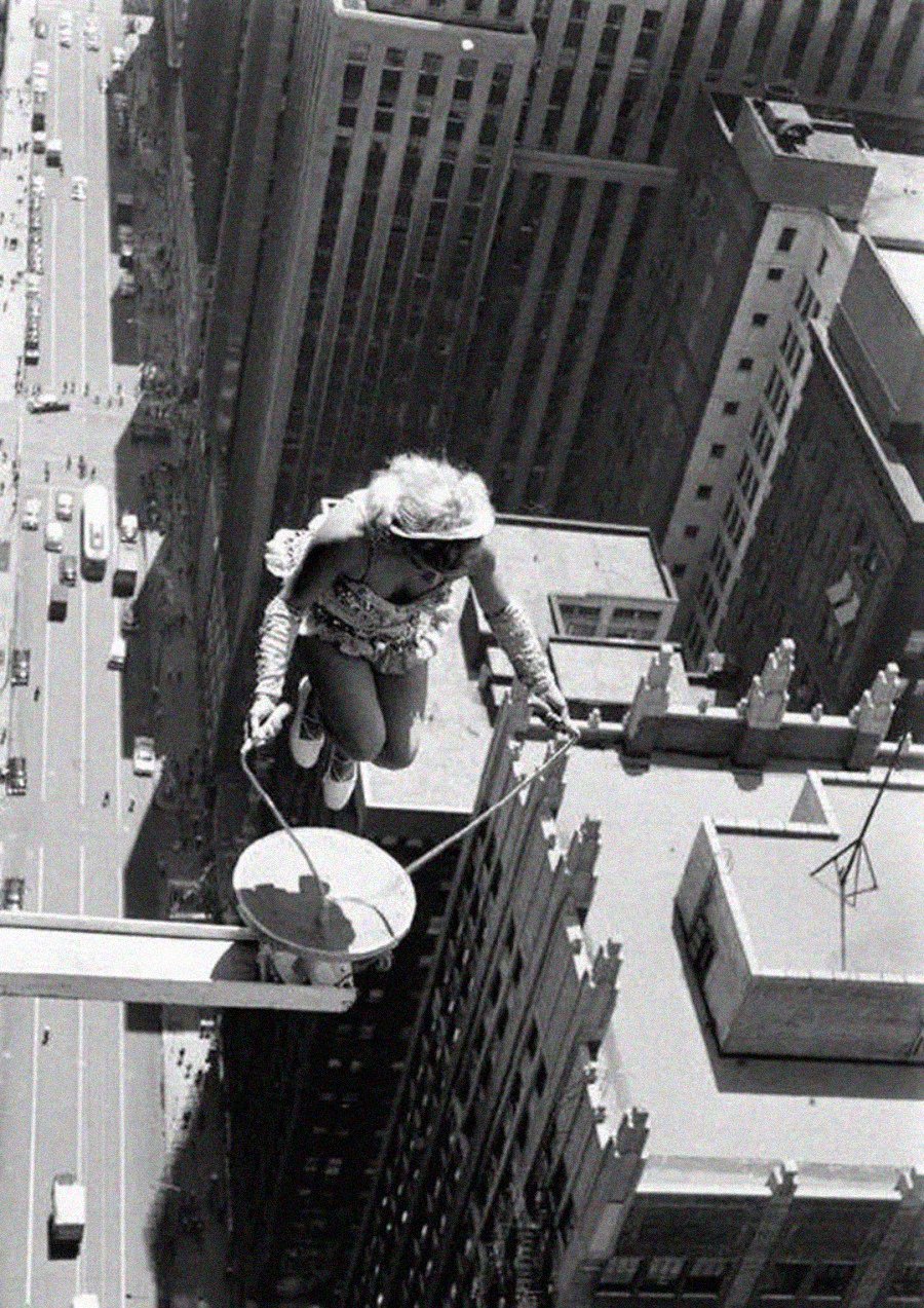 A woman in costume jumps rope on a narrow platform high above a city street, with tall buildings and traffic far below, performing a daring stunt.