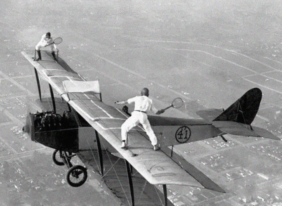 Two people in white outfits play tennis on the wings of a biplane flying high above a landscape, performing a daring stunt mid-air with rackets and a net.