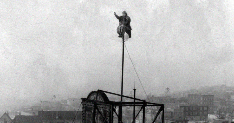 A person balances high atop a narrow pole on a rooftop, overlooking a foggy cityscape filled with buildings. The scene appears daring and dangerous.