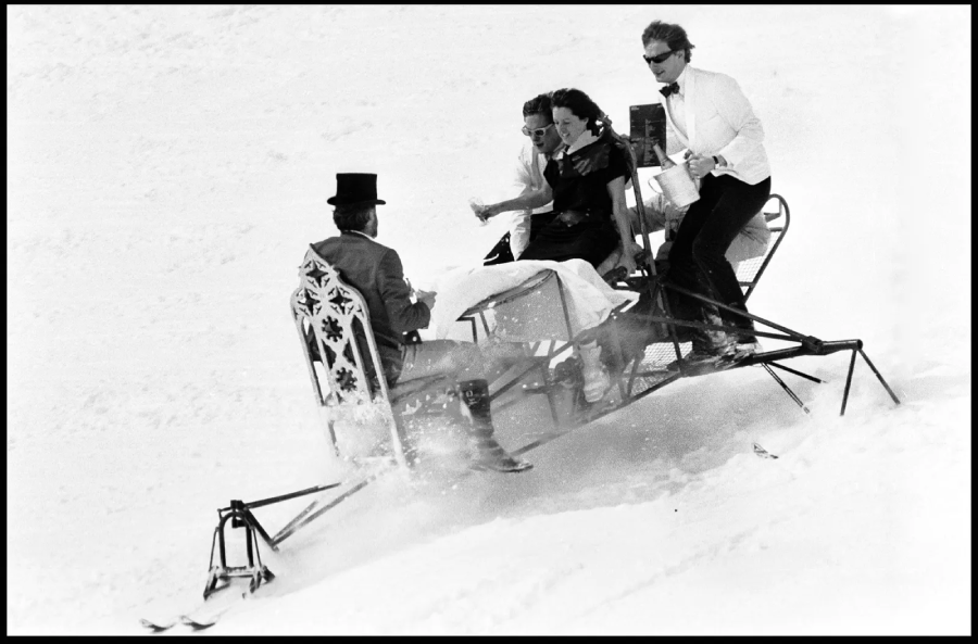 Three people in formal attire ride a decorative sled on snow, with one person in a top hat steering and the others seated behind, all appearing joyful and playful.