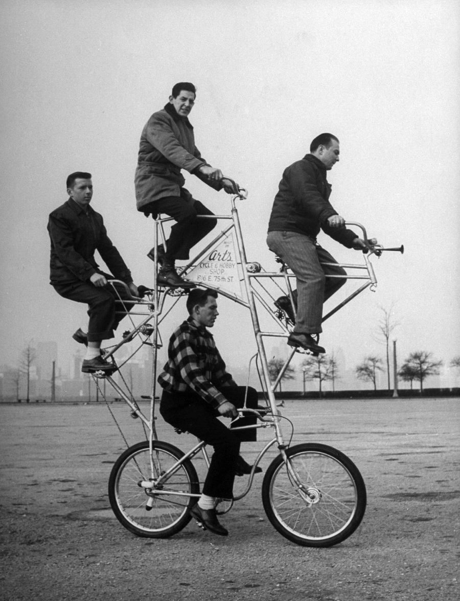 Four men ride a tall, double-decker bicycle. Two men are seated on the lower bike, and two are seated above them on a second bike frame. They are outdoors on a paved area with trees in the background.