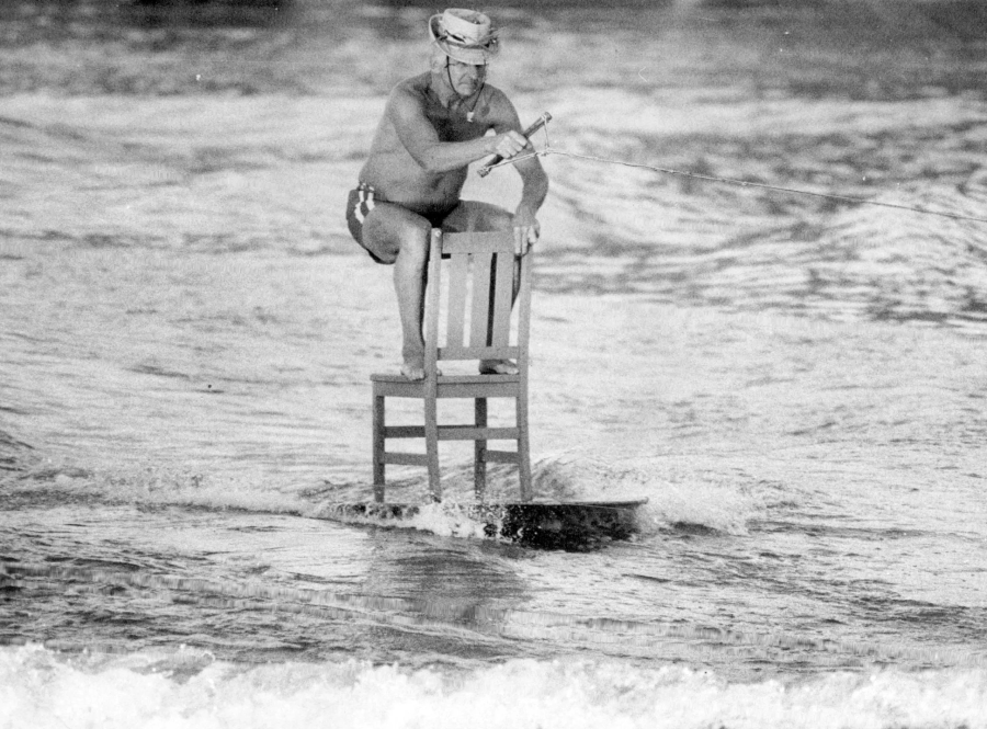 A man wearing a helmet and swim trunks rides a wooden chair placed on a surfboard, holding onto a tow rope while skimming across the water.