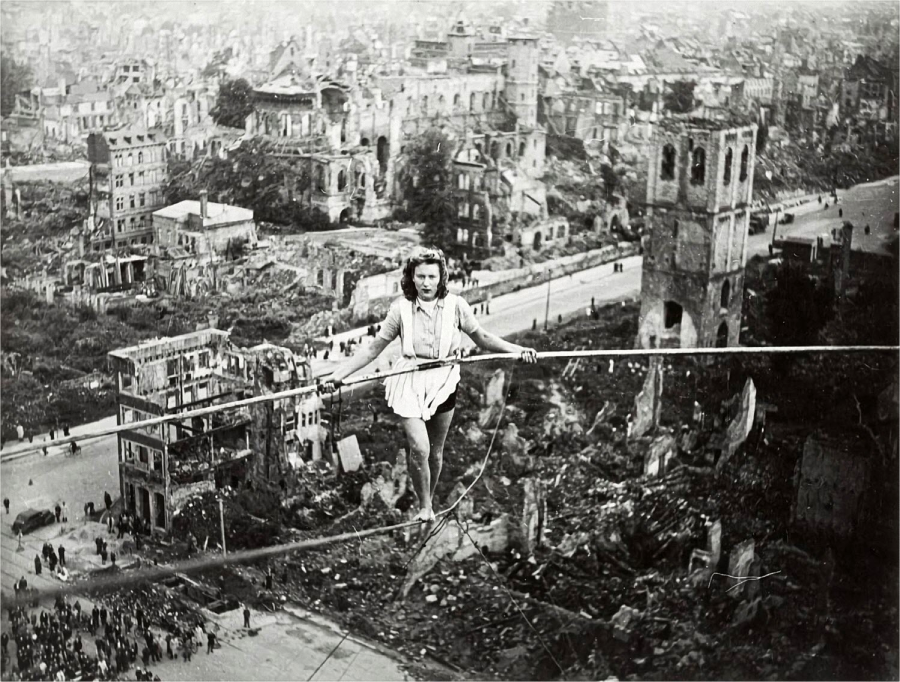 A woman performs a daring tightrope walk high above the ruins of a bombed city, surrounded by destroyed buildings and rubble, with crowds watching from below.