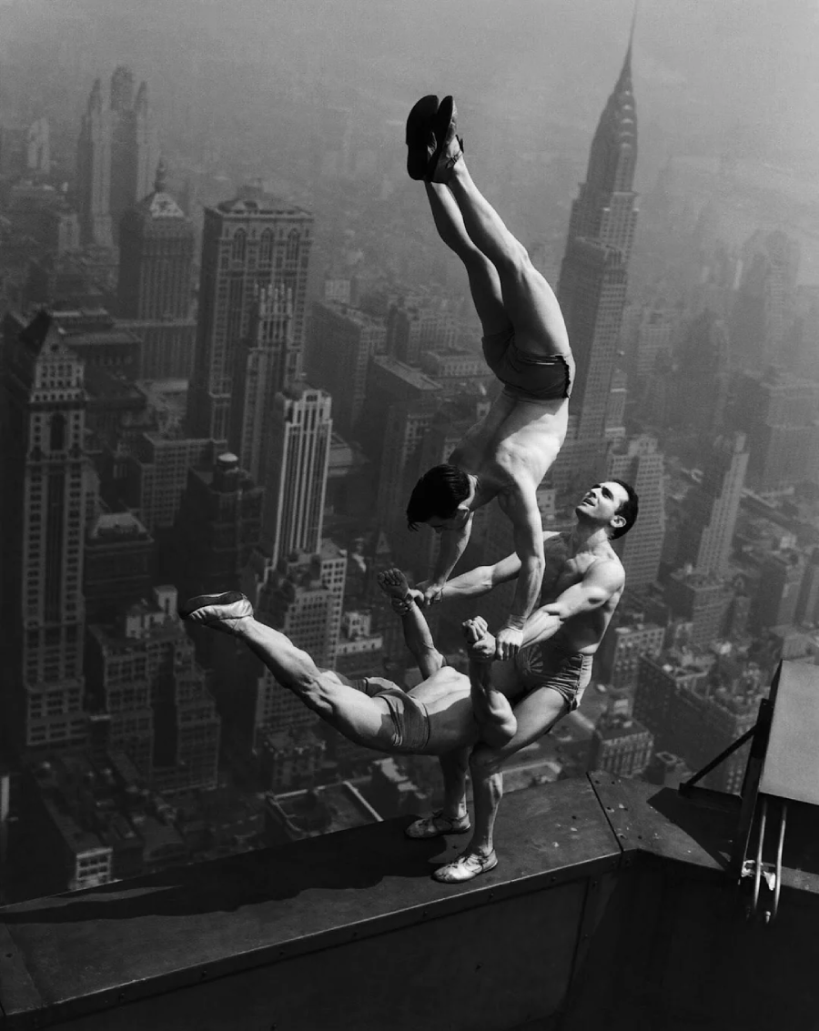 Three acrobats perform a balancing act on the edge of a skyscraper rooftop overlooking a cityscape, with tall buildings, including the Chrysler Building, visible in the background.