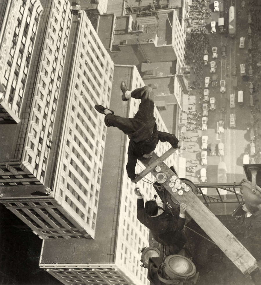 Two men balance on a narrow plank high above city streets, with one man appearing to fall off the edge while the other sits beside a stack of plates, skyscrapers and a crowded street far below.