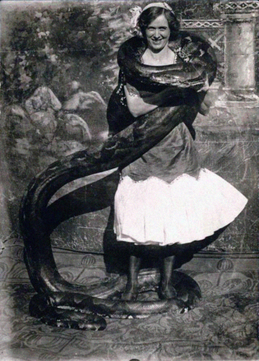 A woman in a skirt and blouse stands smiling as a large snake wraps around her shoulders and legs, set against a painted backdrop.