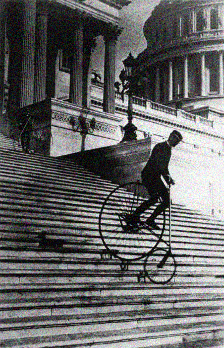 A man rides a vintage penny-farthing bicycle down the stone steps of a grand building with large columns and a domed roof in the background.