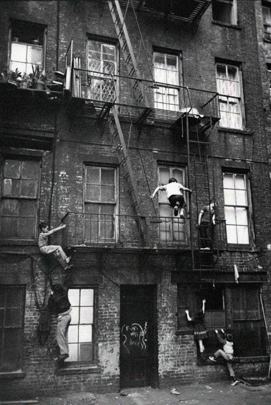 A black-and-white photo shows several children climbing the metal fire escape of an old brick apartment building, with some hanging from ladders and others standing on platforms or clinging to window bars.
