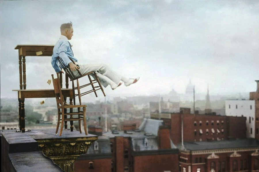 A man in white pants and a blue shirt sits precariously on a backward-tilted chair, stacked atop more furniture, teetering on the edge of a tall building with a cityscape in the background.