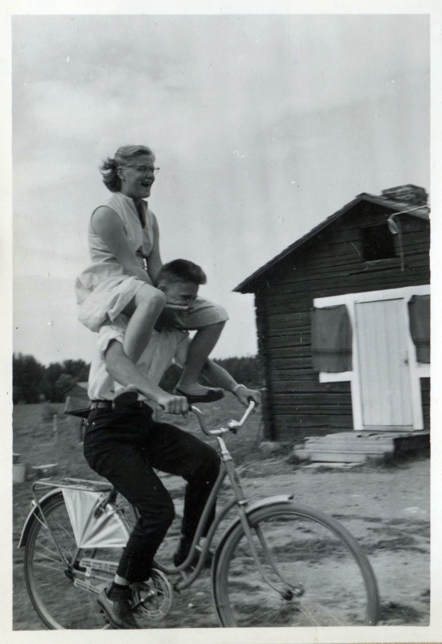 A man rides a bicycle with a woman sitting on his shoulders. They appear to be outdoors near a wooden building. The photo is black and white, and both are smiling, creating a playful, joyful scene.