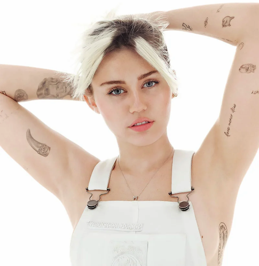 A person with short, blonde hair poses with their arms above their head, revealing various tattoos on both arms. They are wearing white overalls and looking directly at the camera against a bright white background.