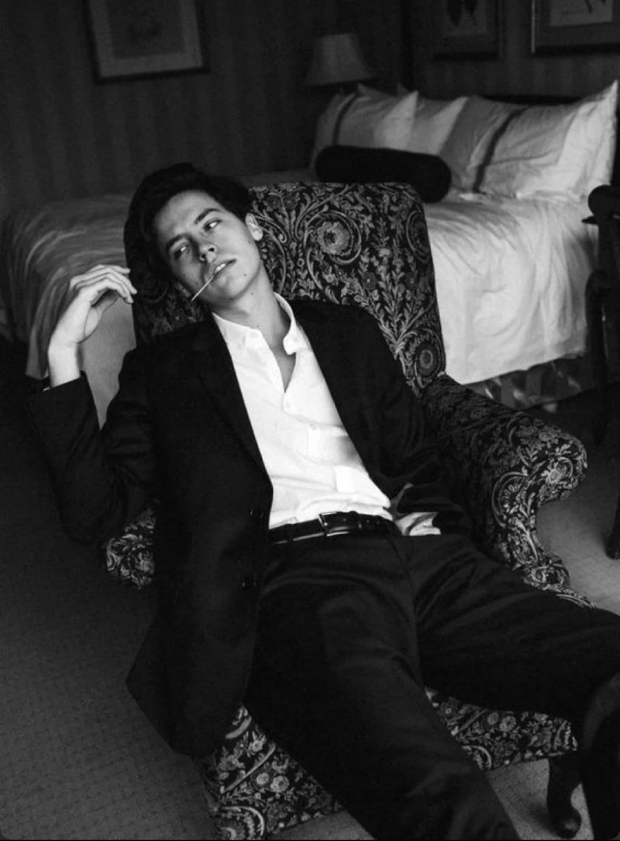 A person in a suit sits slouched in a patterned armchair, holding a cigarette, with a relaxed pose in a dimly lit bedroom featuring an unmade bed in the background. The photo is in black and white.