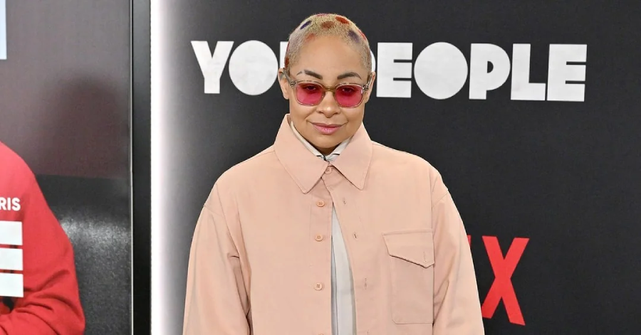 A person with short, light-colored hair and red designs stands in front of a "You People" Netflix backdrop, wearing pink-tinted sunglasses and a light pink jacket over a light shirt.