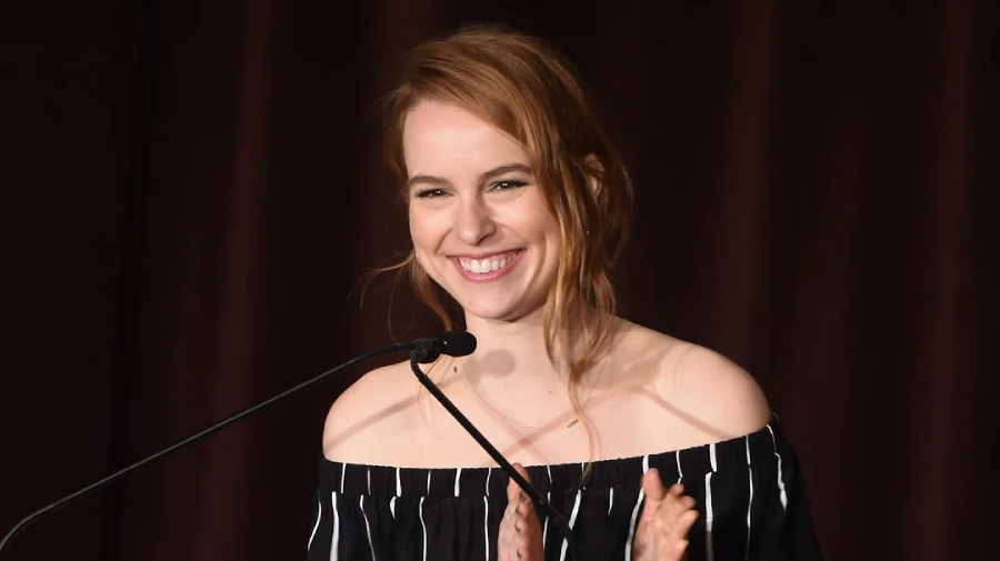 A young woman with long, light brown hair smiles while speaking at a microphone. She is wearing an off-the-shoulder black top with white vertical stripes, and the background is dark and out of focus.