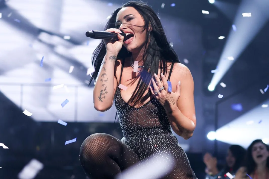 A singer with long dark hair passionately performs on stage, holding a microphone and kneeling amidst falling confetti. She wears a sparkly, sleeveless outfit and fishnet stockings, with audience members visible in the background.