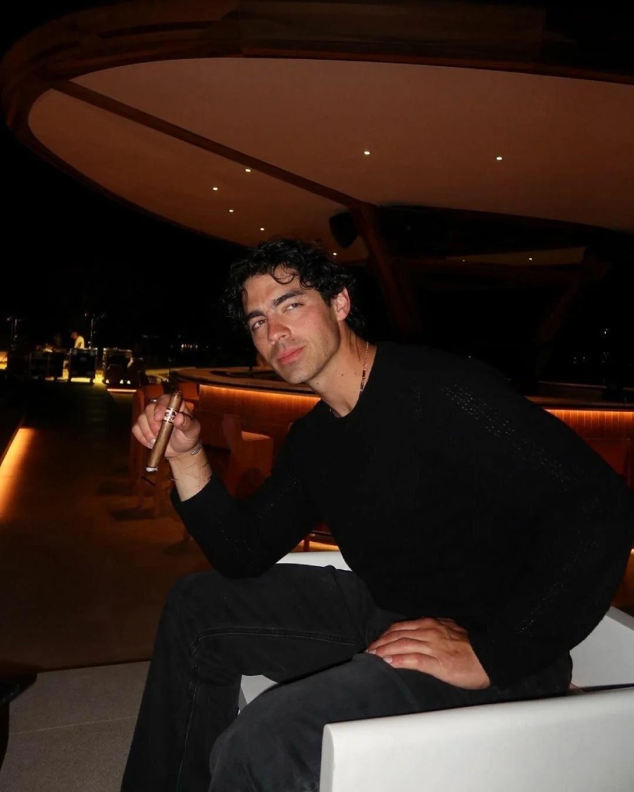 A man with curly dark hair, wearing a black long-sleeve shirt and dark pants, sits on a white chair at a dimly lit modern bar at night, holding a cigar in his right hand and looking toward the camera.
