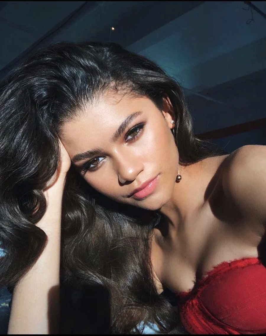 A woman with long, wavy dark hair and glowing makeup poses for a selfie in natural sunlight, wearing a strapless red top and looking confidently at the camera.