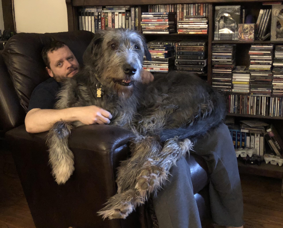 A man sits in a recliner with a large, shaggy grey dog sprawled across his lap. Shelves filled with DVDs and CDs are visible in the background.