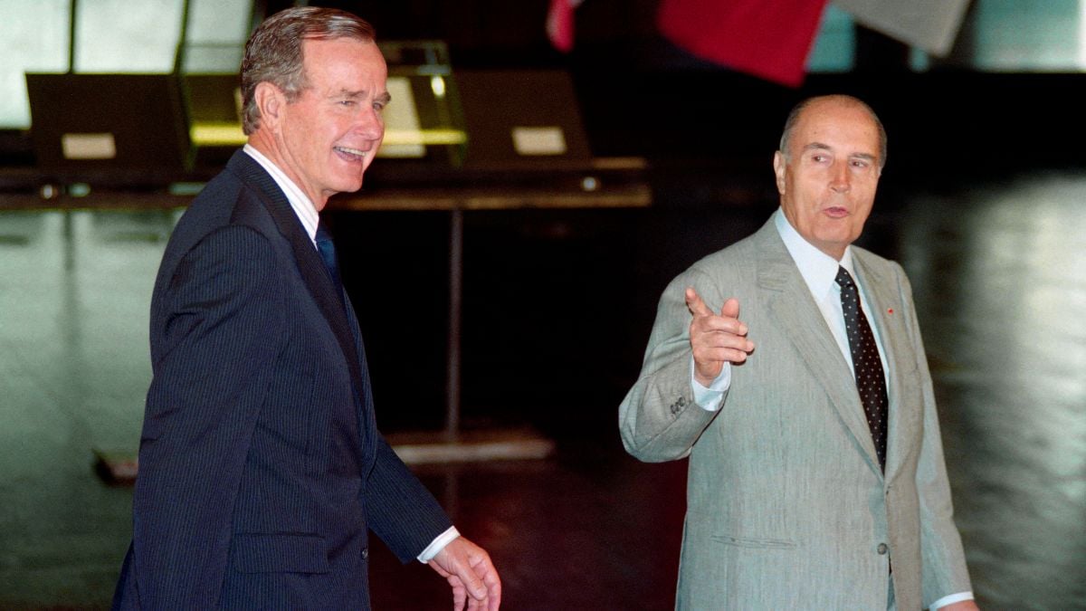 Two men in suits converse indoors; one is smiling while the other gestures with his hand. They appear to be engaged in a formal or diplomatic setting.