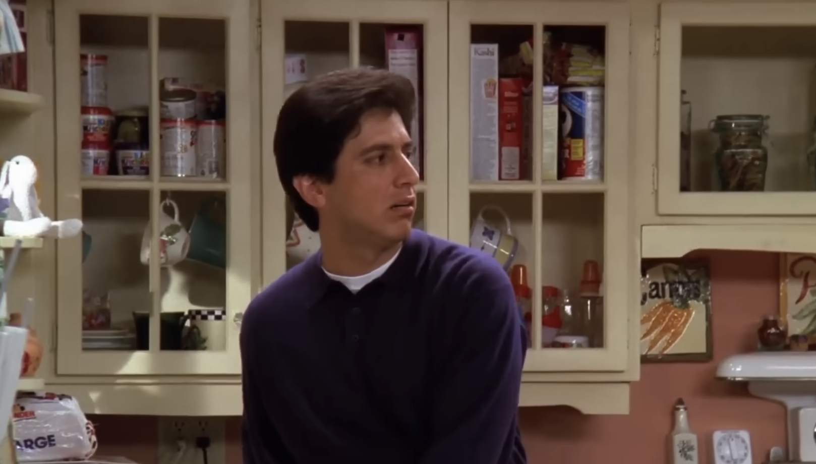 A man with dark hair wearing a navy blue shirt sits in a kitchen, turned to his left, looking concerned. Behind him are shelves with food items and kitchen supplies.