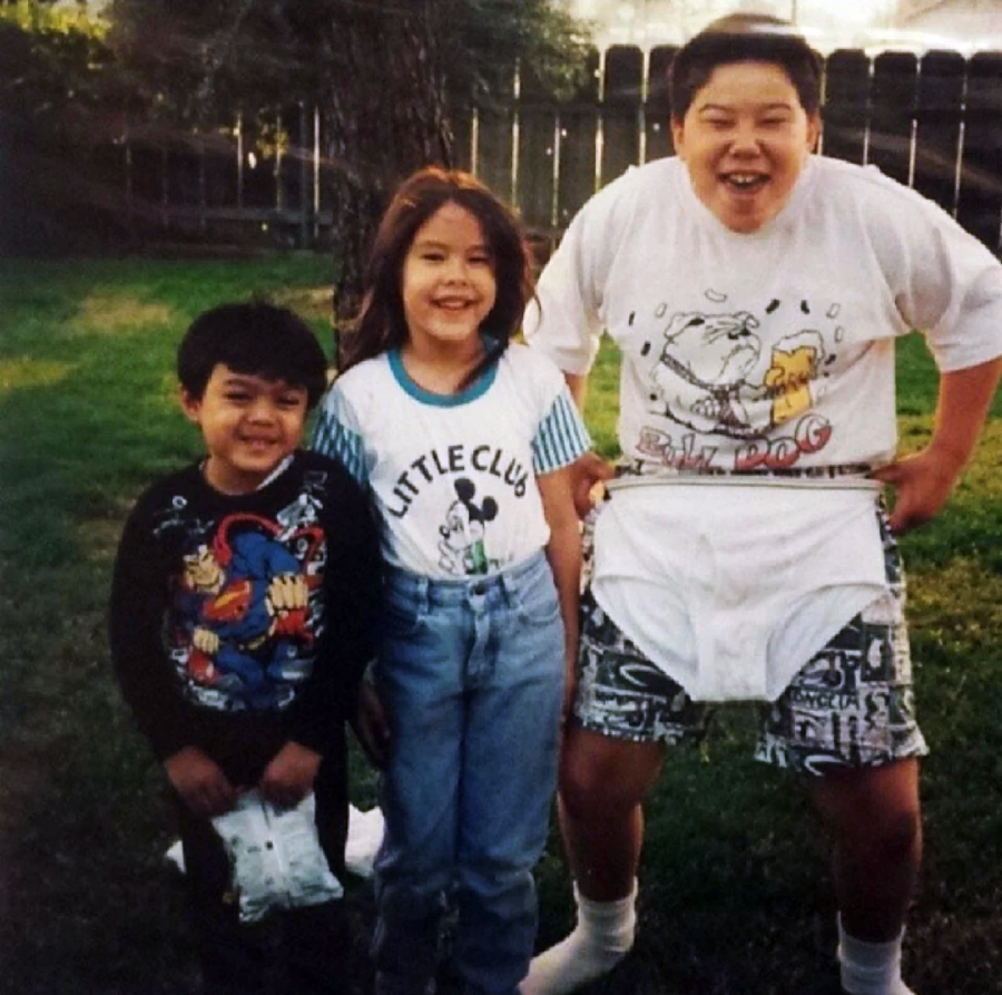 Three children are standing on grass in a backyard, smiling at the camera. The oldest child on the right is wearing large cartoon underwear over his shorts, while the other two children stand close together, also smiling.