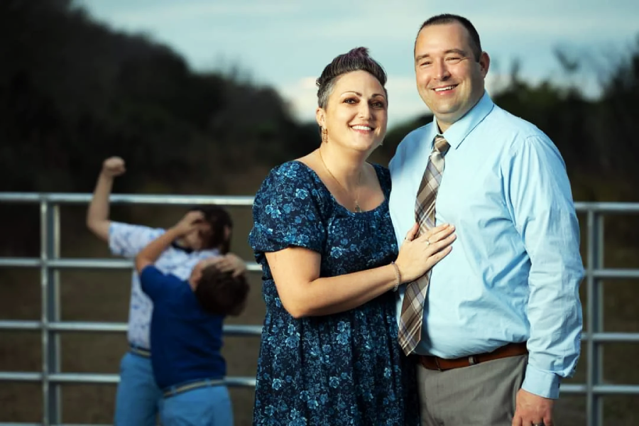 A smiling couple poses for a photo in front of a fence, while two boys playfully wrestle in the background, with trees and a cloudy sky behind them.