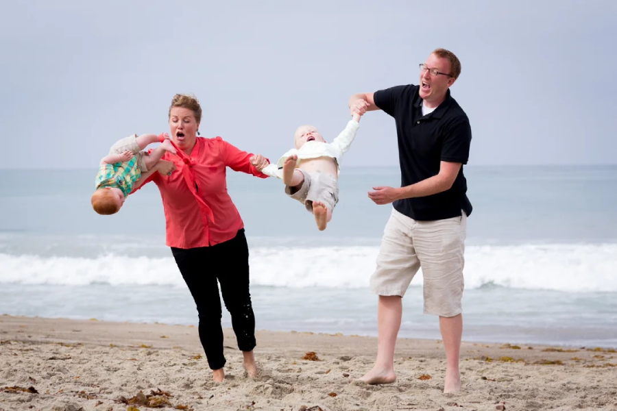 A woman and man on a beach each swing a baby by the arms, the babies lifted off the ground and smiling. The adults look surprised and playful, with the ocean and waves in the background.