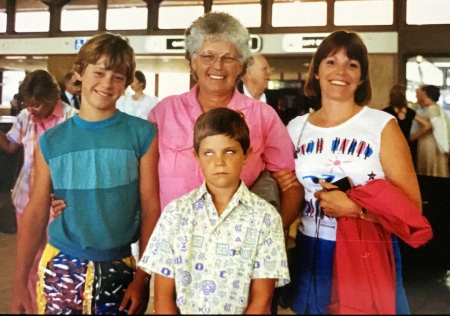 Three women and one young boy pose for a photo at what appears to be an airport. The boy is standing in front and rolling his eyes upward, while the others are smiling at the camera. There are people and signs in the background.