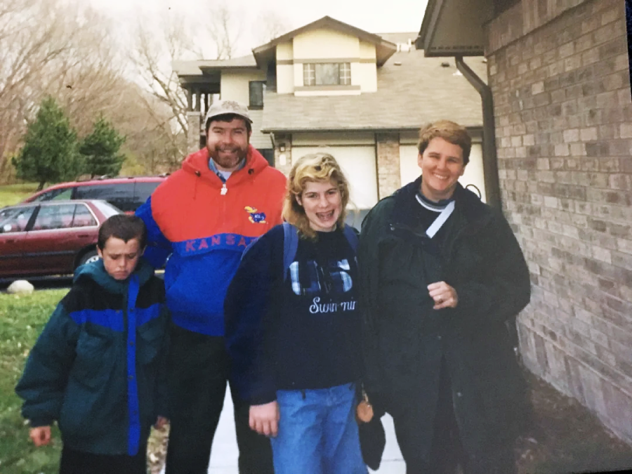 Four people stand outside near a brick house. Two adults smile at the camera, one wearing a red and blue jacket. A teenage girl in a sweatshirt grins, while a young boy in a coat looks down, not smiling. A red car is in the background.