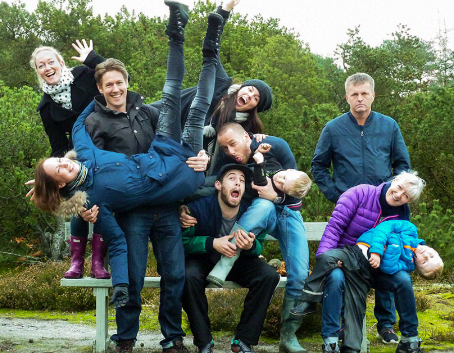 A group of ten people, adults and children, pose playfully outdoors; some are lifting others, making funny faces, or sitting on a bench. One person stands off to the side, looking serious. Trees and greenery are in the background.