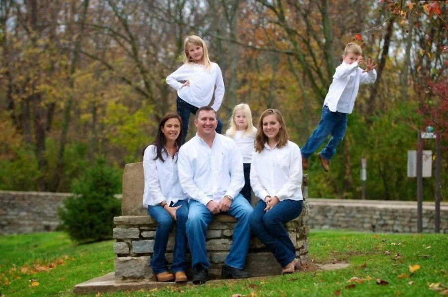 A family of six, all wearing white shirts and jeans, poses outdoors on a stone bench. Five sit or stand together while one child is captured mid-air, jumping to the right. Autumn trees fill the background.