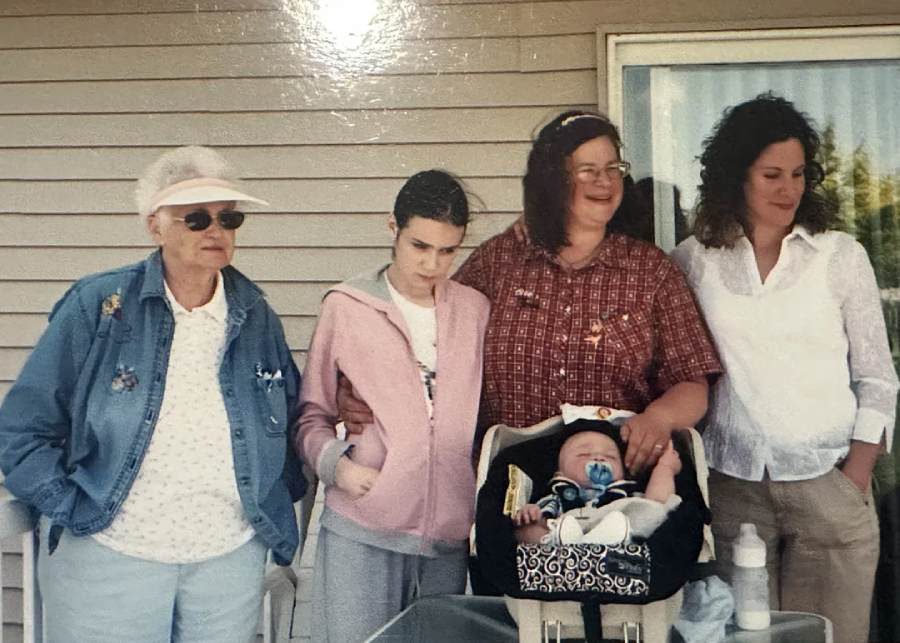Five people stand in front of a tan paneled wall. An elderly woman in a visor, a girl in pink, and two women stand behind a baby in a car seat. A baby bottle is on the table beside them.
