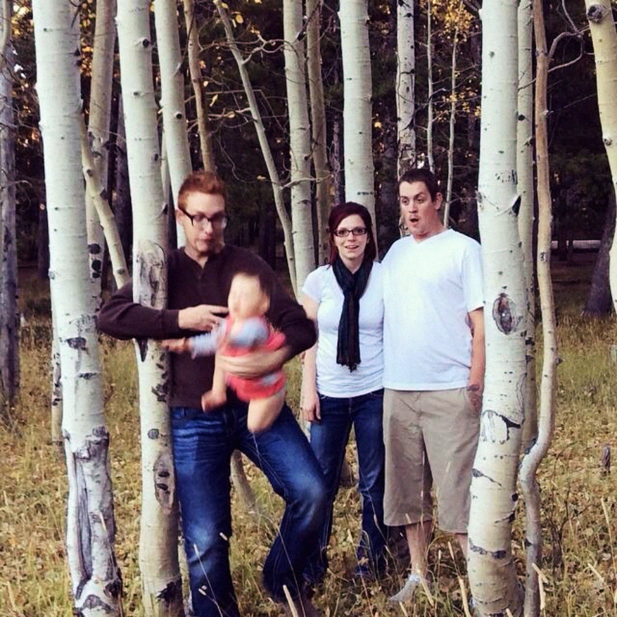 Three adults stand among aspen trees in a forest. One man in the foreground is swinging a blurred baby, while the other two adults, a woman and a man, stand behind them, both looking surprised.