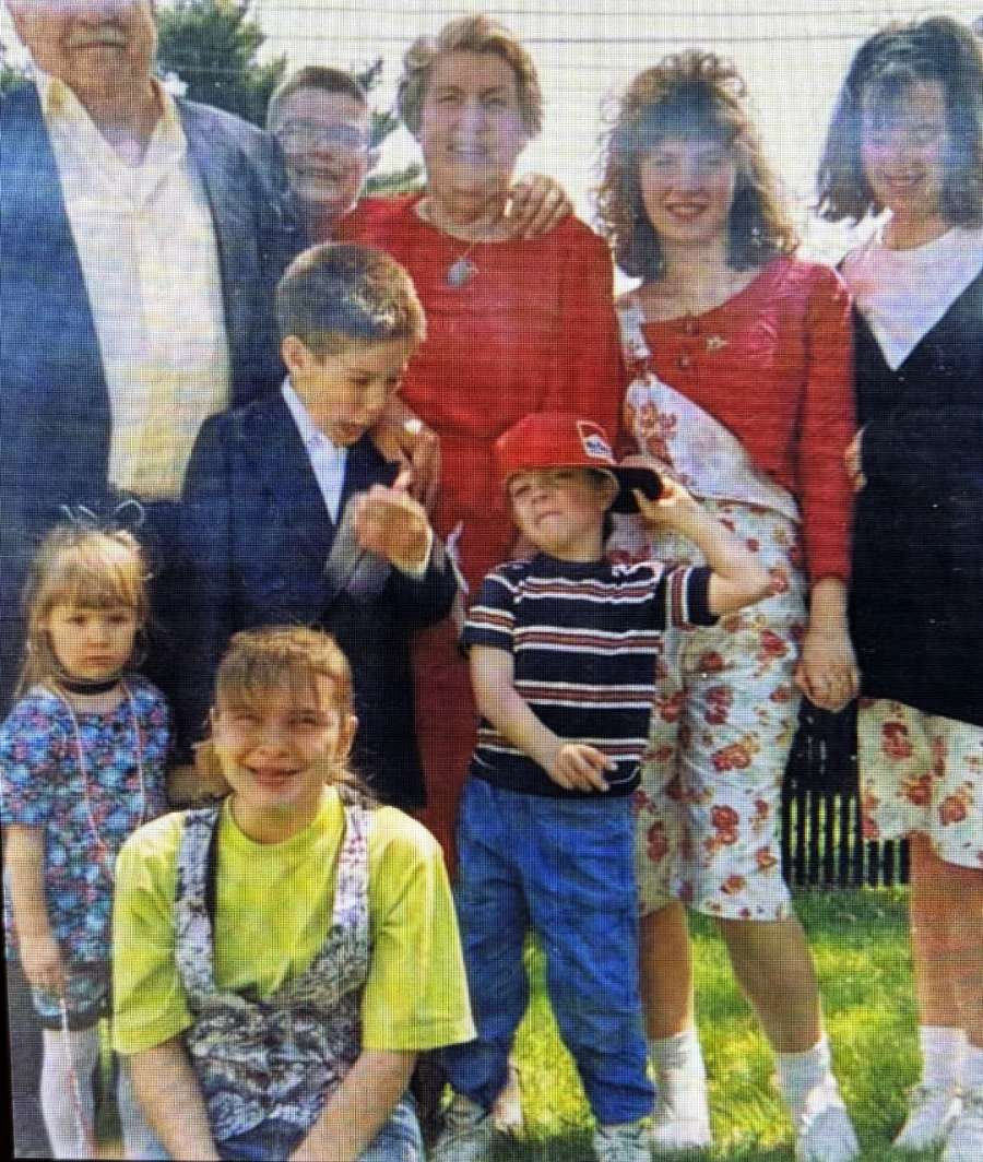 A group of people, including adults and children, pose together outdoors on grass. Most are smiling, and the sun is shining. One child in a red cap strikes a playful pose while others stand, with trees and a fence in the background.