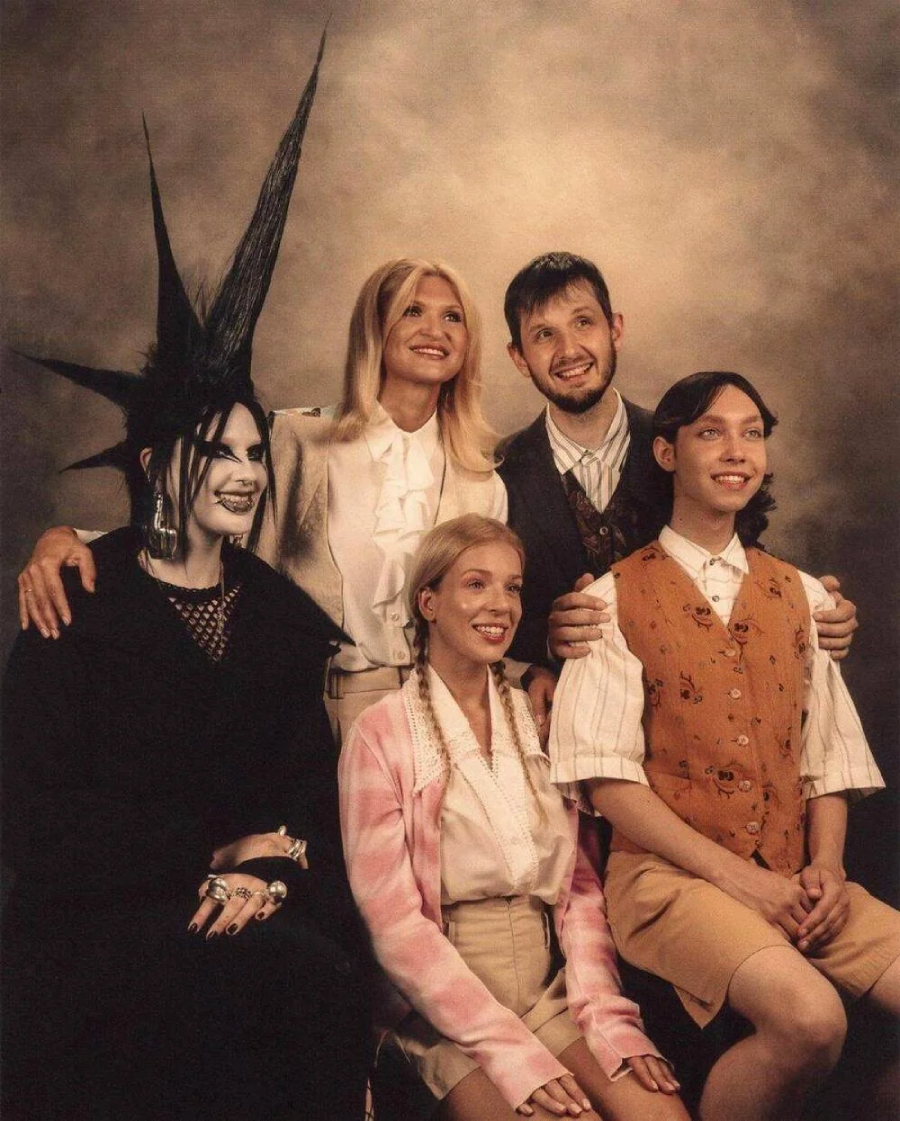 A group of five people pose for a vintage-style family portrait. Four are smiling. One person, dressed in black with dramatic spiked hair and makeup, contrasts with the others, who wear light, retro-inspired outfits.