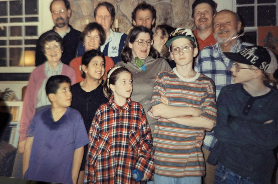 A group of thirteen people, including adults and children, pose closely together indoors, some smiling and others making playful expressions, with a stone wall and windows in the background.