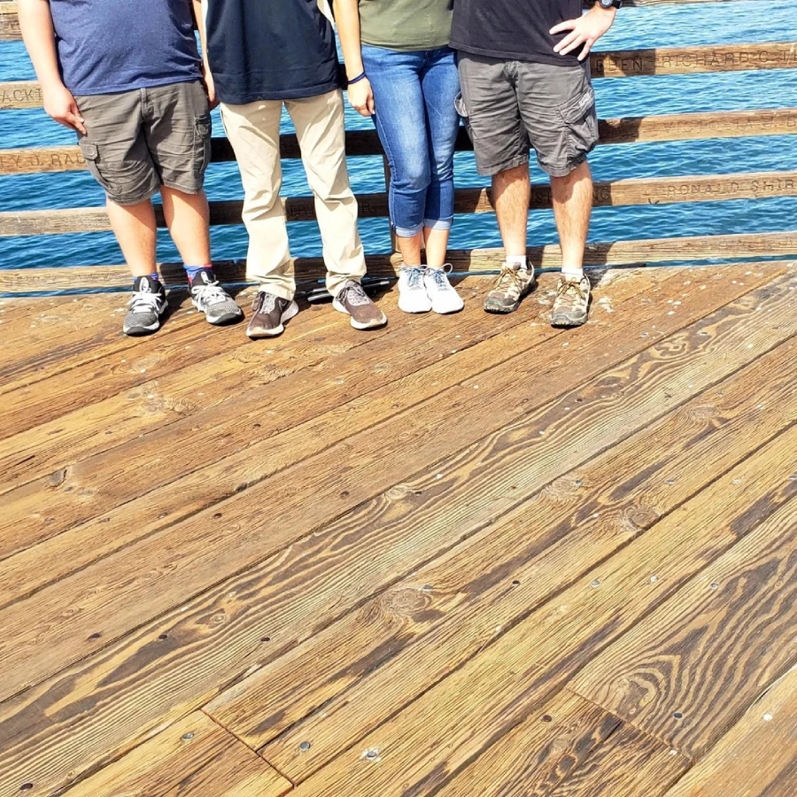 Four people stand side by side on a wooden pier with water in the background. The photo is cropped at their shoulders, showing only their bodies from the neck down.