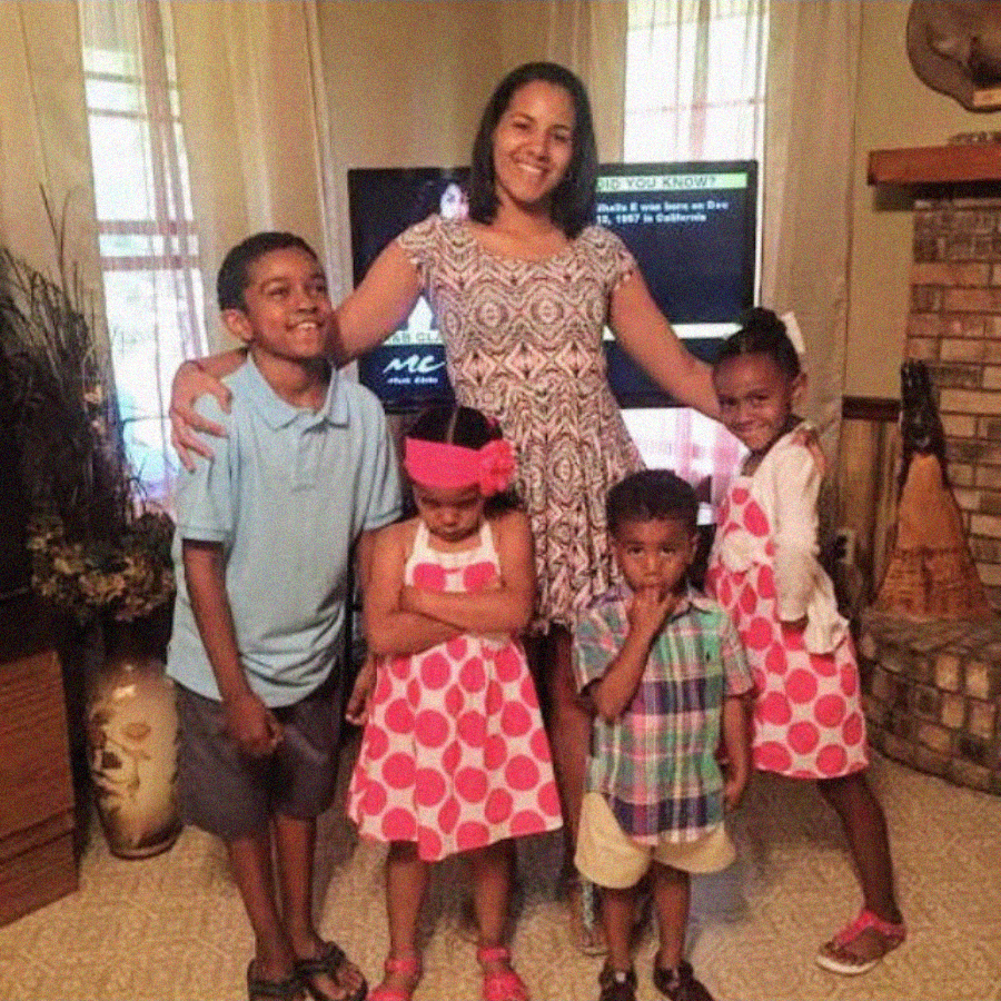 A woman stands indoors with four young children. Two girls in matching pink polka dot dresses, a boy in a blue shirt, and a younger boy in plaid stand in front of her. Everyone is smiling except one girl with arms crossed.