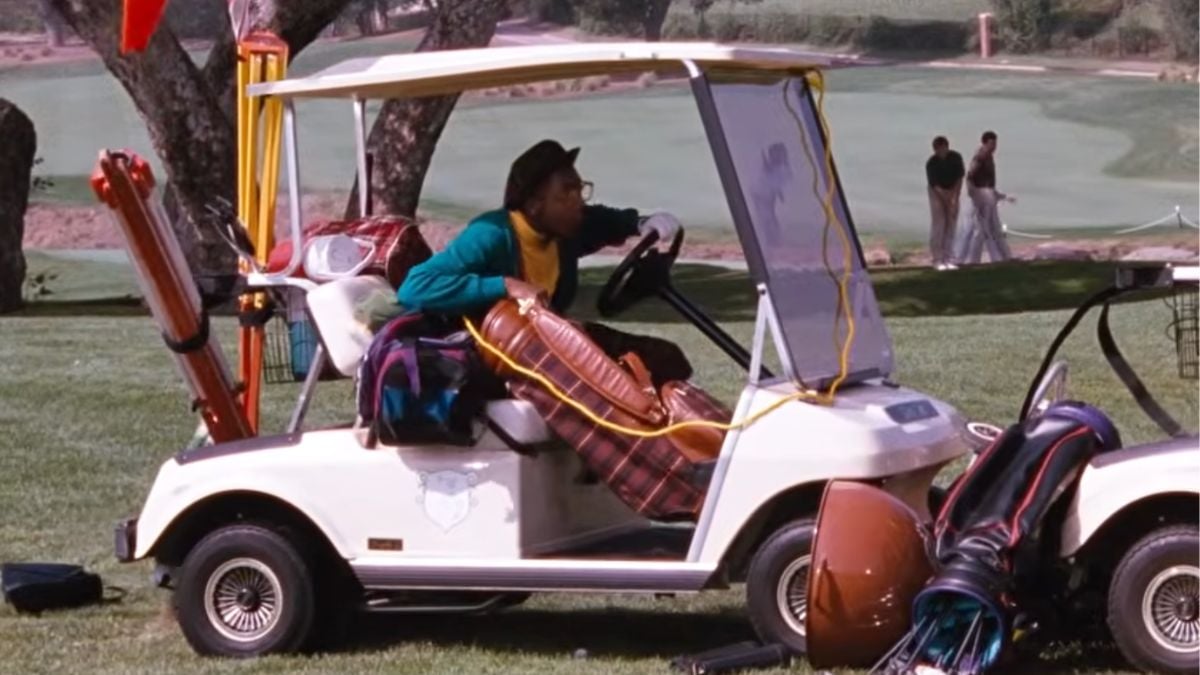 A person sits in a golf cart filled with golf bags and equipment on a grassy course. Two people in formal attire stand in the background near the edge of the course.