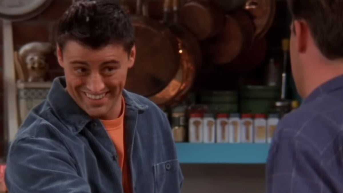 A man with dark hair smiles while wearing a blue jacket and orange shirt, standing in a kitchen with spice jars and hanging pots in the background. Another person is partly visible, facing him.