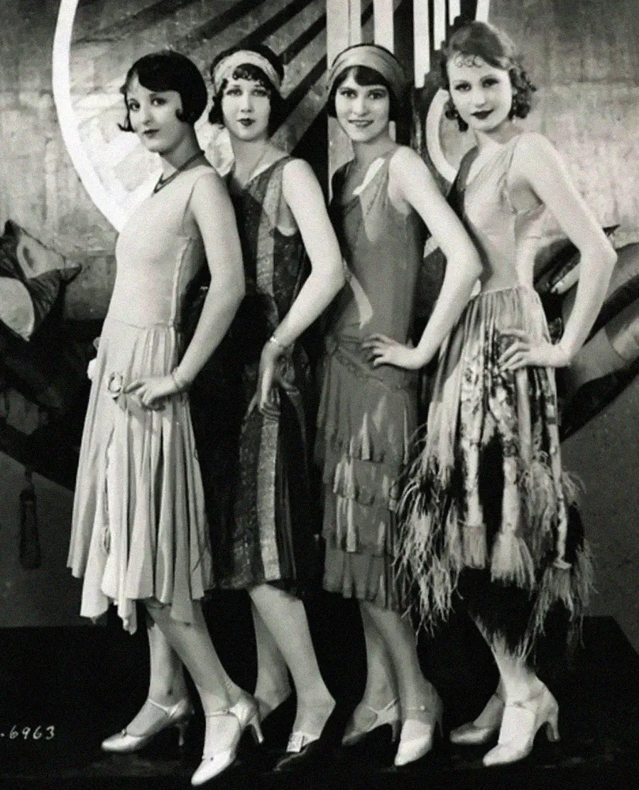 Four women in 1920s flapper dresses pose side by side, each with short, styled hair and hand on hip, standing in front of an art deco backdrop.