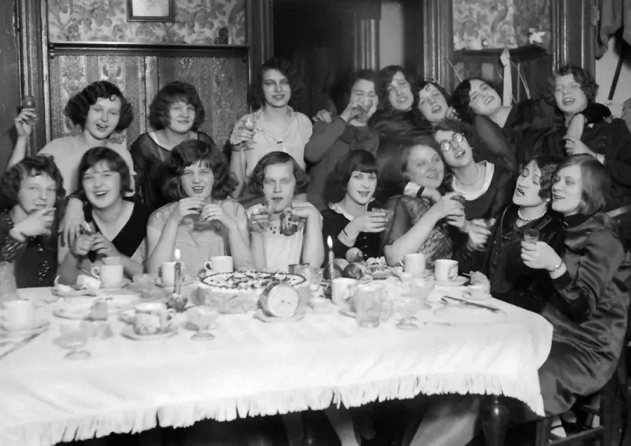 A group of women in 1920s-style clothing gather around a table set with teacups and a cake, smiling and raising glasses in celebration inside a decorated room.
