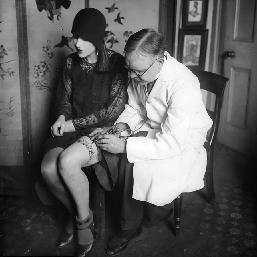A woman in 1920s attire sits while a man in a white coat tattoos a design on her knee. They are indoors, with patterned wallpaper and framed pictures on the wall behind them.