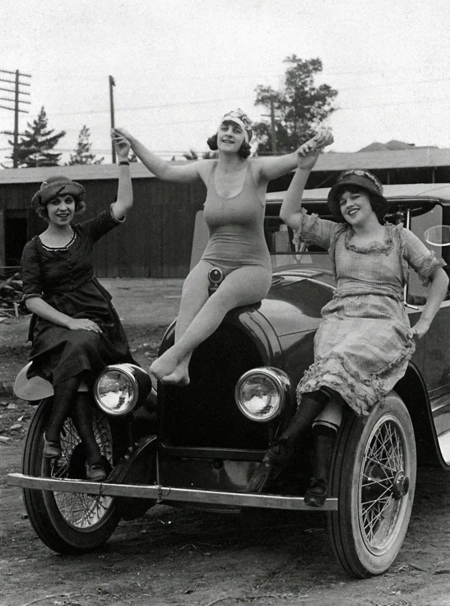 Three women from the early 20th century smile and pose on the hood of a vintage car; one in a swimsuit sits in the center, while the other two in dresses sit on either side, all holding hands and laughing.
