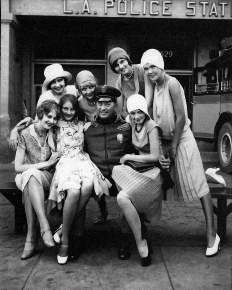 A group of smiling women in 1920s flapper dresses pose with a uniformed police officer on a bench outside an L.A. police station; a vintage car is parked nearby.