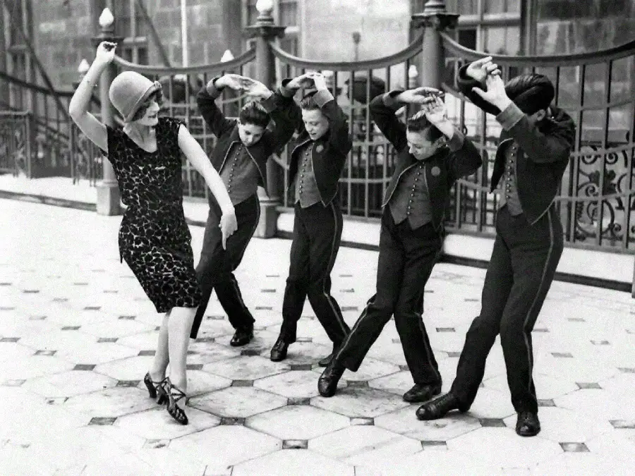A woman in a patterned dress and cloche hat dances energetically on a tiled street while three boys in matching uniforms mimic her movements, arms raised above their heads. Iron railings and a building appear in the background.
