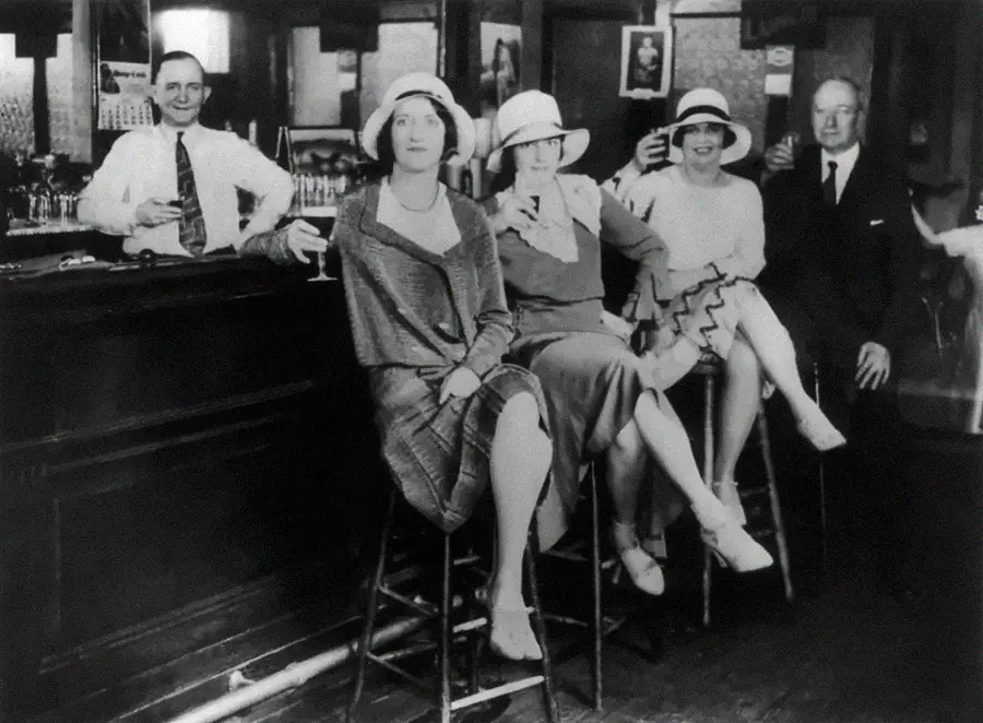 Black and white photo of three women in 1920s dresses and hats sitting on stools at a bar, flanked by two men in suits, all holding drinks and smiling. The setting appears lively and social.