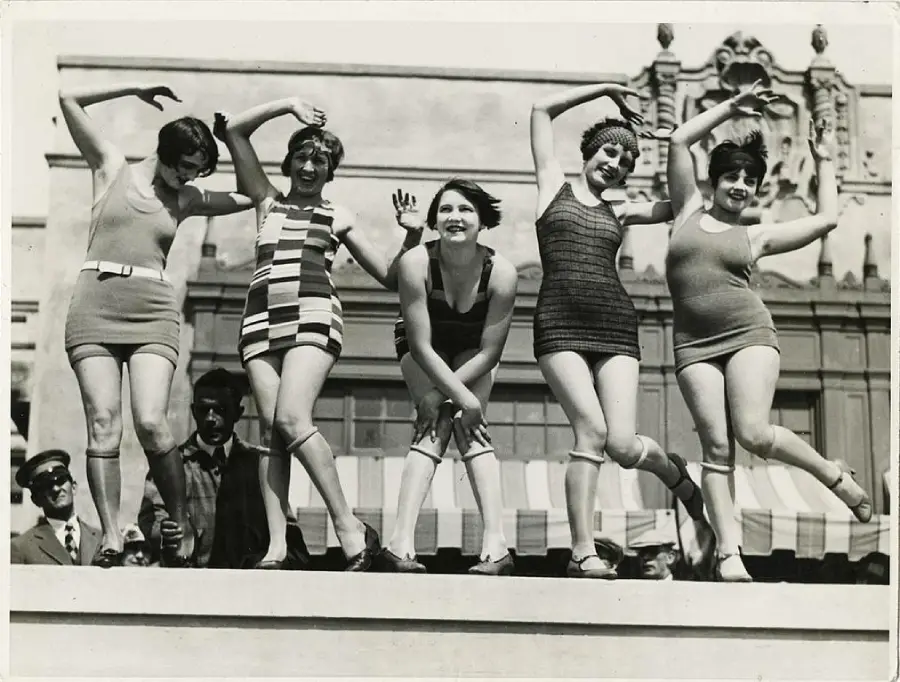 Five women in vintage swimsuits pose playfully on a platform, smiling and raising their arms. The background shows an ornate building and striped awnings, with onlookers below. The image is black and white.