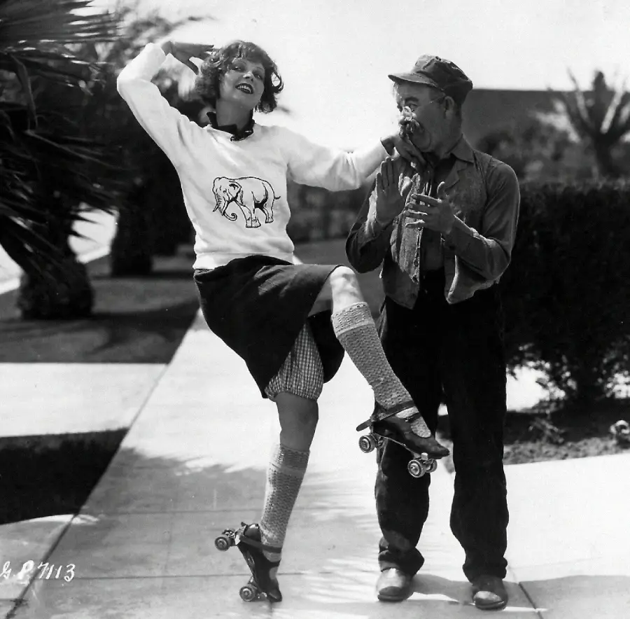 A woman in a skirt and roller skates lifts her leg playfully near a surprised man. She wears a sweater with an elephant design and long socks. They are outdoors on a sidewalk with trees in the background.