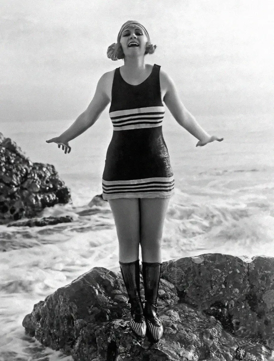 A woman in a vintage swimsuit and swim cap stands on a rock by the sea, smiling with arms outstretched. Waves crash in the background in this black-and-white photo.